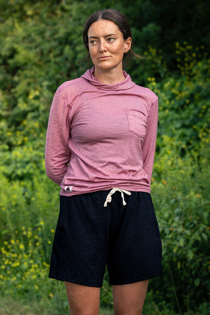 A woman standing outdoors in front of a green foliage background, wearing a dusty rose long-sleeve athletic top with a chest pocket and dark charcoal shorts with a white drawstring.