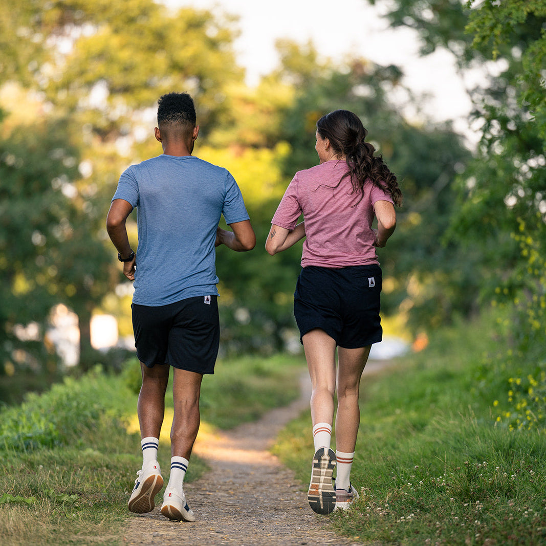 A man and a woman jogging from behind on a dirt path surrounded by green trees and grass, wearing athletic apparel.
