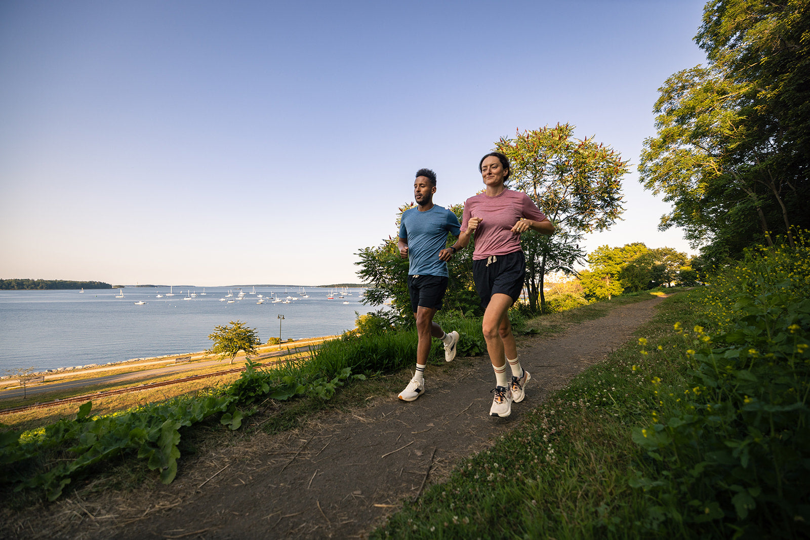 Two people jogging side by side on a dirt path next to the ocean with boats in the distance, surrounded by green trees and grass.
