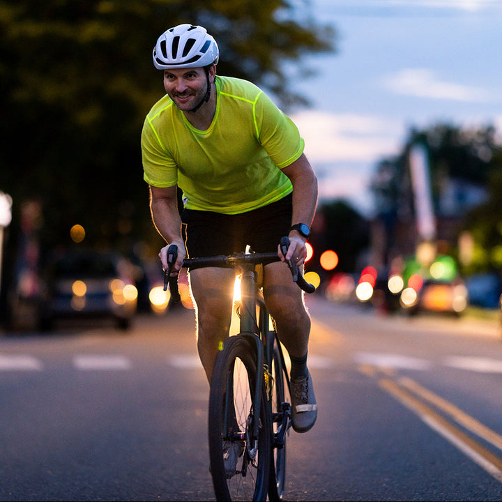 A man wearing a white bicycle helmet, a bright neon yellow-green t-shirt, and black shorts rides a road bike on a city street at dusk.