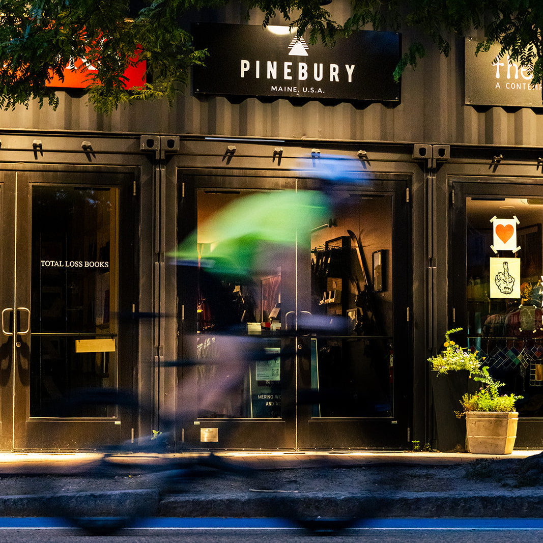 Pinebury Handmade storefront at dusk with a blurred cyclist in front of the glass doors and a tree branch hanging over the entrance.