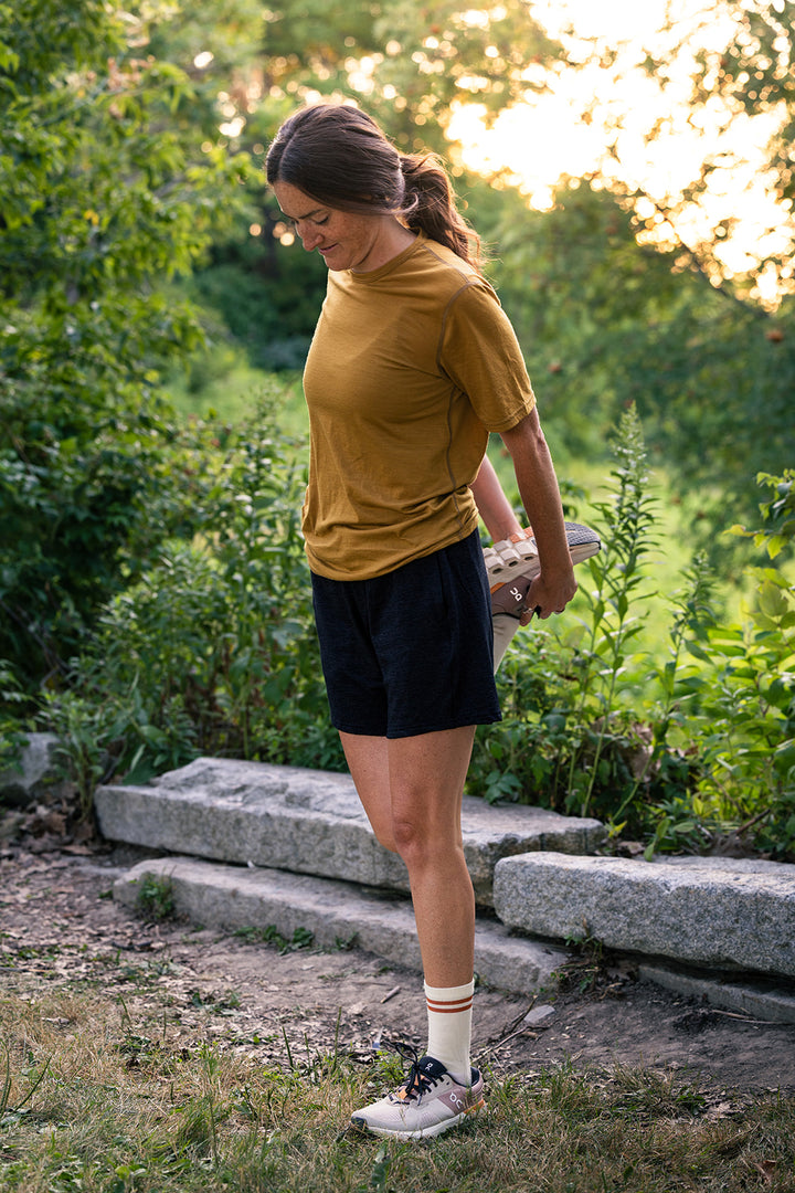 A woman standing outdoors in a wooded setting, wearing a mustard yellow short-sleeve athletic T-shirt, black athletic shorts, white crew socks, and white sneakers, holding a small book or notebook under her arm.