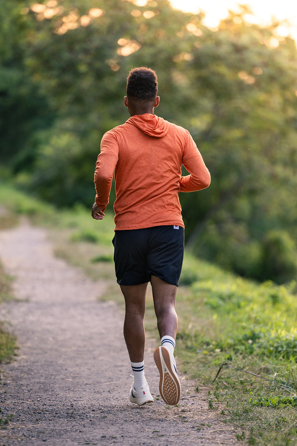 A man running on a dirt trail in a forest, viewed from behind. He is wearing an orange long-sleeve hooded athletic top, black running shorts, white socks, and running shoes.