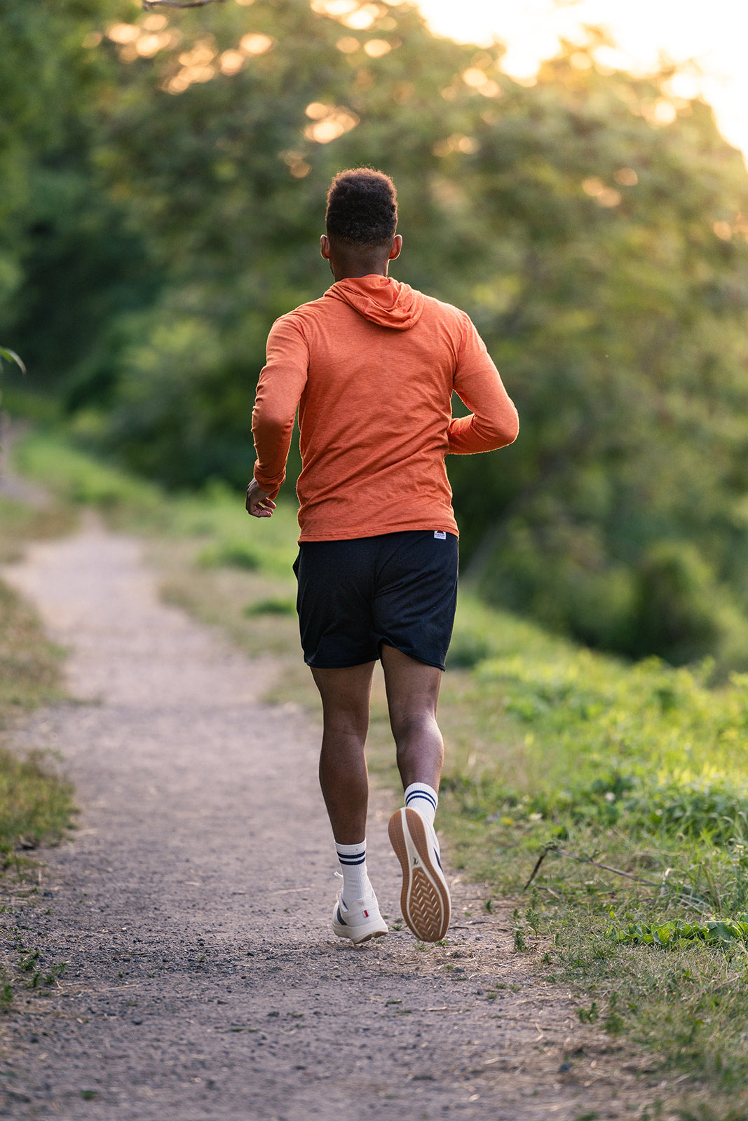A man running on a dirt trail in a forest, viewed from behind. He is wearing an orange long-sleeve hooded athletic top, black running shorts, white socks, and running shoes.