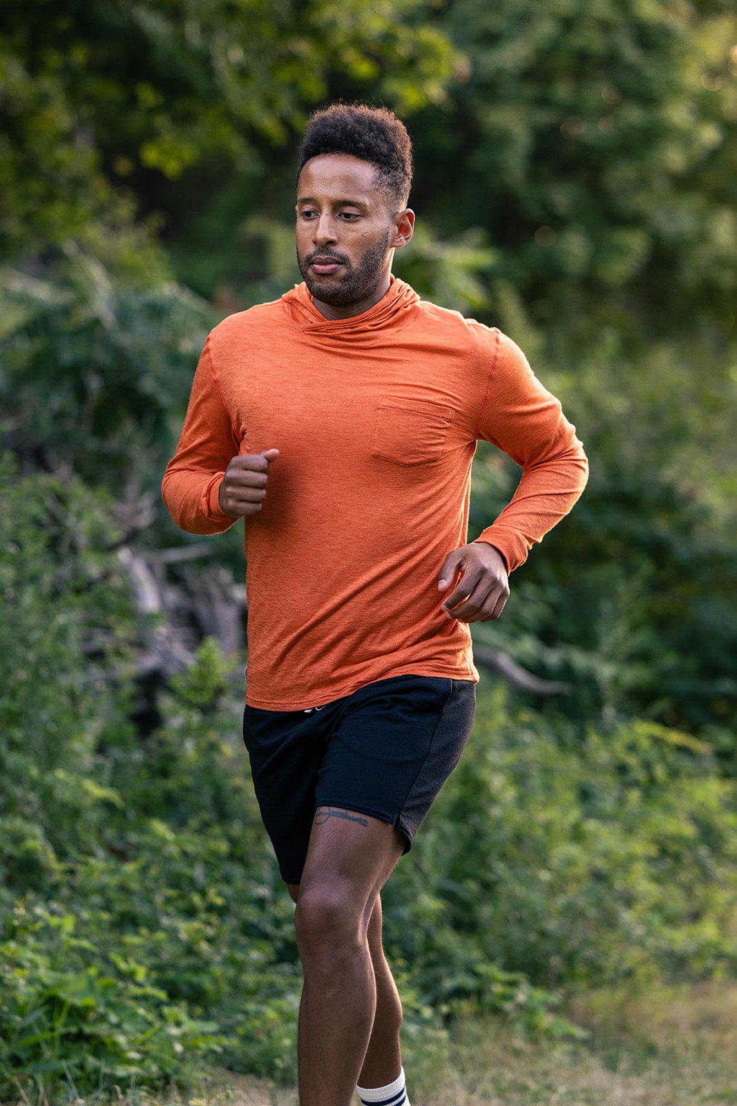 A man jogging outdoors wearing a bright orange long-sleeve athletic turtleneck top and black running shorts, with green foliage in the background.