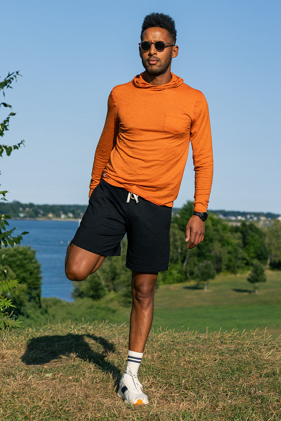 A man wearing an orange long-sleeve athletic hoodie and black athletic shorts stands outdoors on a grassy hill with a lake and trees in the background.