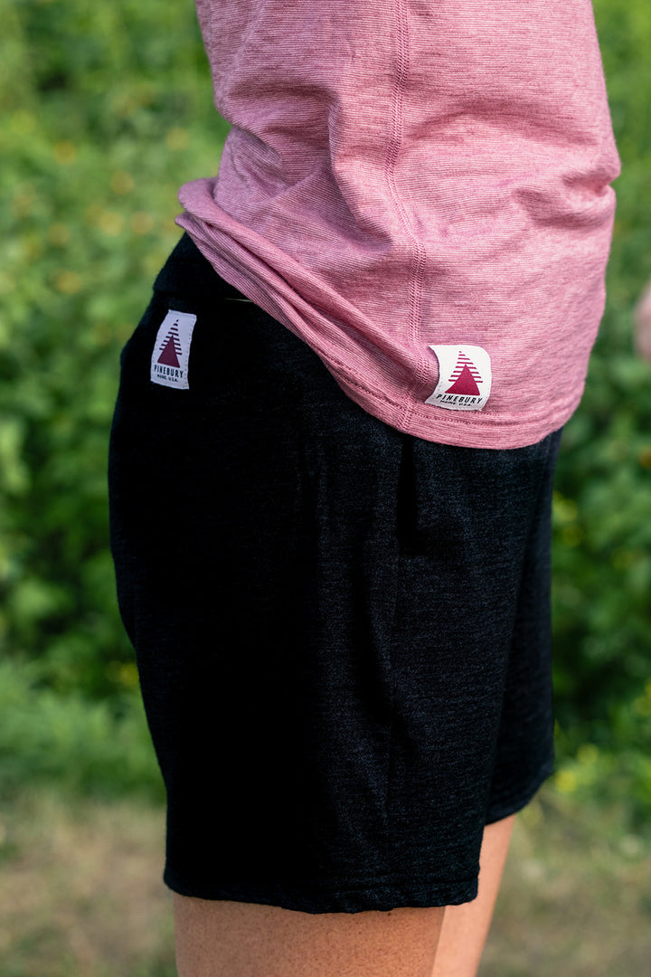 A close-up side view of a person wearing black athletic shorts and a heathered pink t-shirt, with a blurred green outdoor background. The shorts feature a small white brand patch on the side pocket.