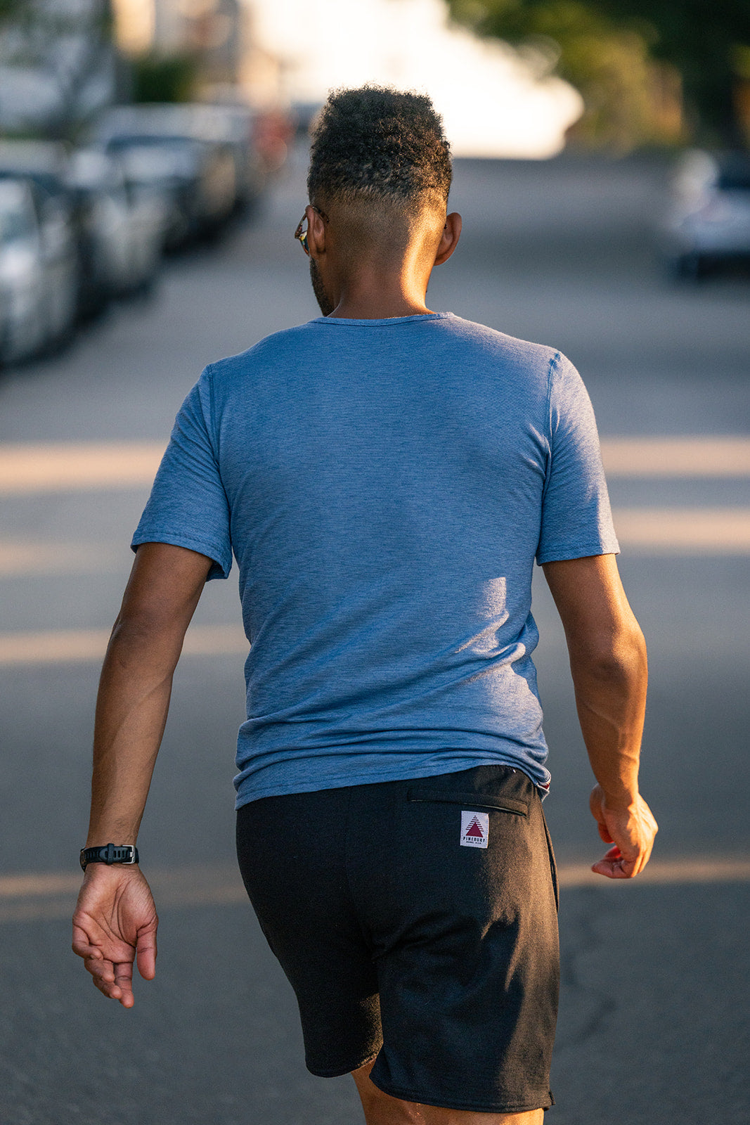 A man viewed from behind walking down a sunlit street, wearing a heathered blue crew-neck t-shirt and black athletic shorts with a small white triangular logo patch on the back pocket.