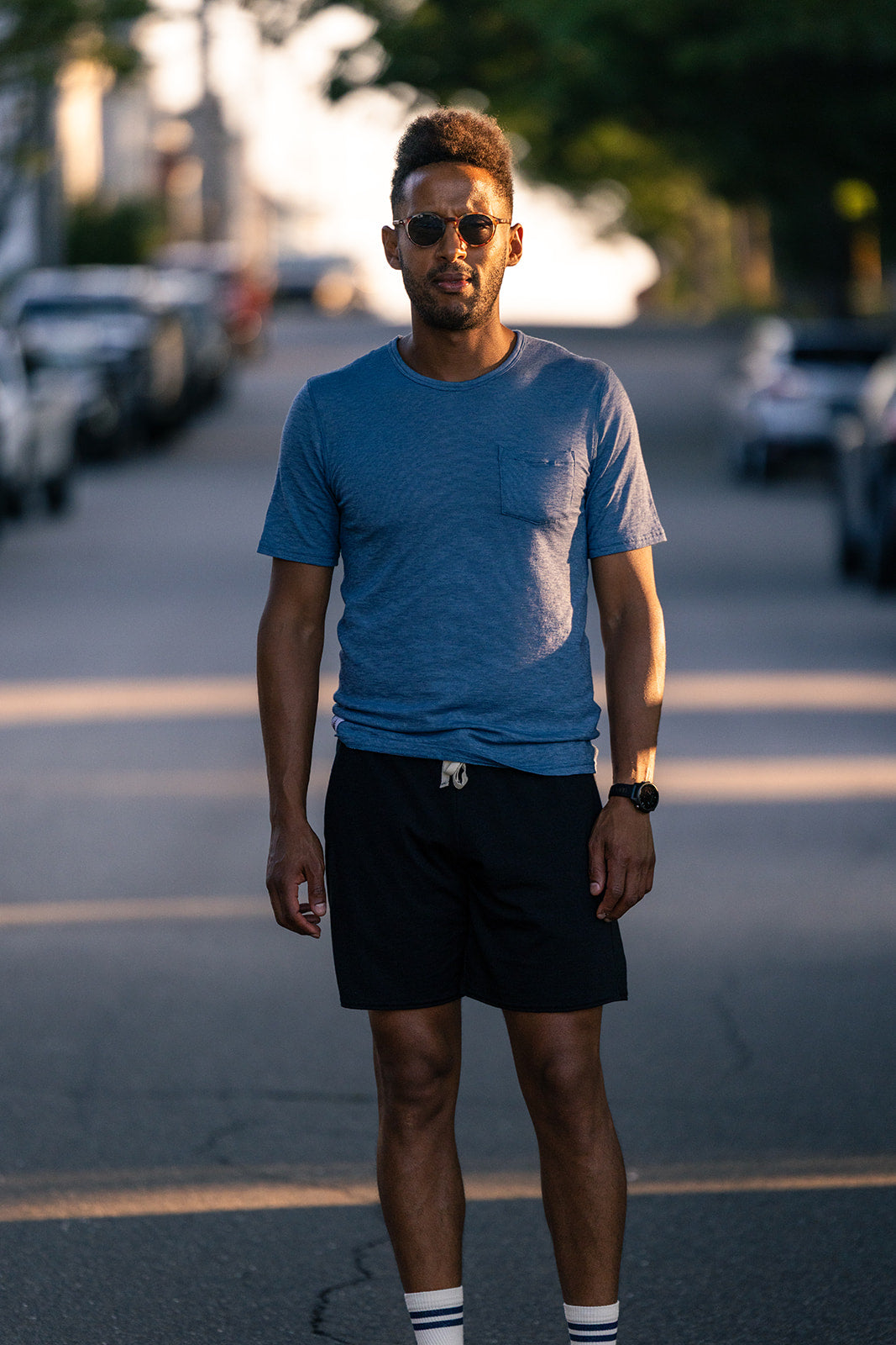 A man wearing a heather blue crewneck t-shirt, black shorts, and sunglasses stands on an asphalt street during golden hour, with parked cars and trees blurred in the background.