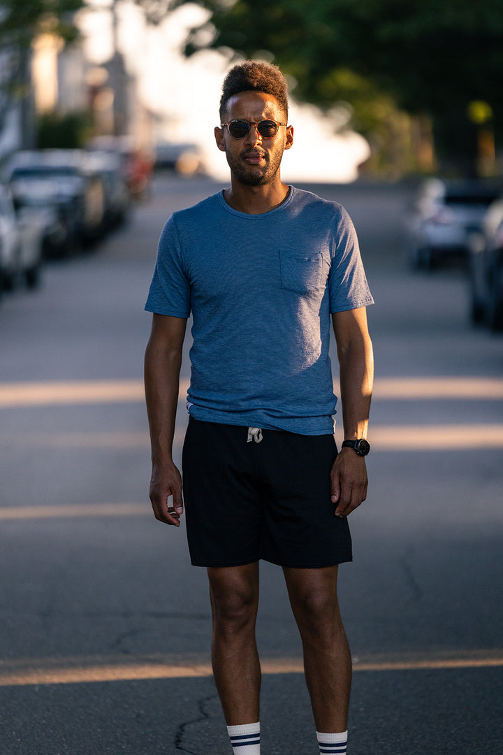 A man wearing a heather blue crewneck t-shirt, black shorts, and sunglasses stands on an asphalt street during golden hour, with parked cars and trees blurred in the background.