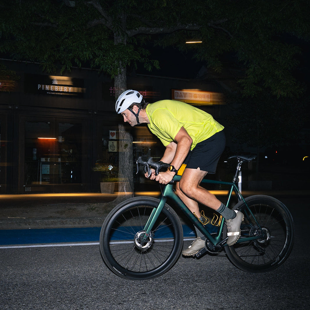 A cyclist wearing a bright yellow-green jersey, black shorts, and a white helmet rides a dark green road/gravel bicycle at night on a street with blurred storefront lights in the background.