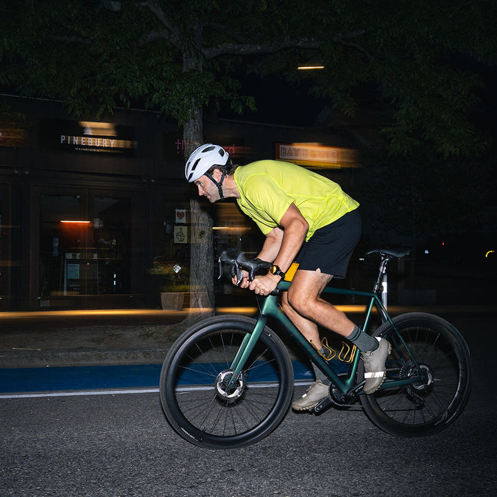 A cyclist wearing a bright yellow-green jersey, black shorts, and a white helmet rides a dark green road/gravel bicycle at night on a street with blurred storefront lights in the background.