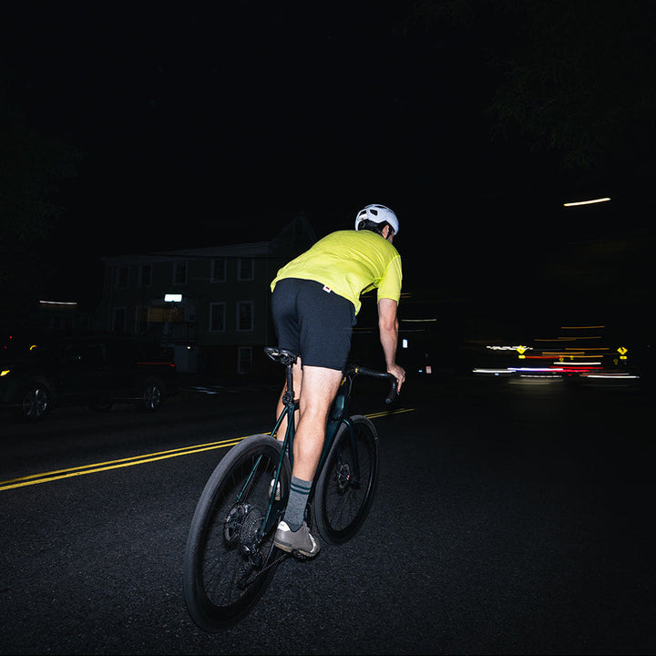 A cyclist rides away on a dark road at night, wearing a bright yellow-green jersey, black shorts, and a white helmet. The scene is very dark with some blurred lights and motion blur on the left side.