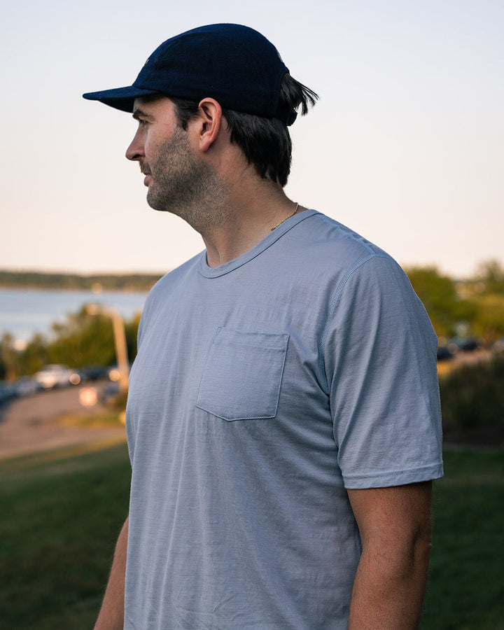 A man wearing a light blue crewneck pocket t-shirt and a dark navy baseball cap, standing outdoors in a grassy area near water during late afternoon.