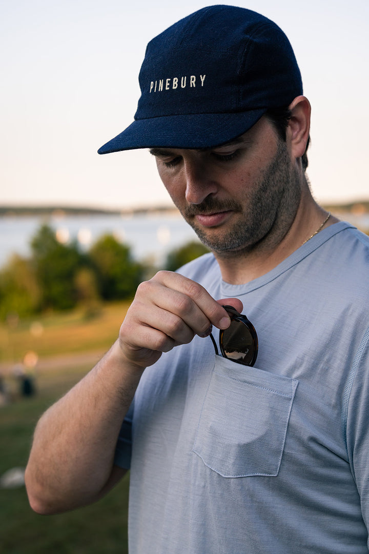 A man wearing a navy blue Pinebury baseball cap and a light blue t-shirt with a chest pocket, holding sunglasses in his hand. The background shows a softly blurred outdoor landscape with water and trees.