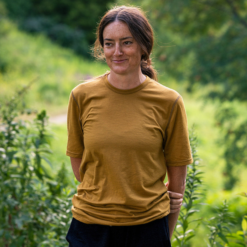 A woman wearing a mustard yellow short-sleeve t-shirt and black athletic shorts, standing outdoors on stone steps with green foliage in the background.