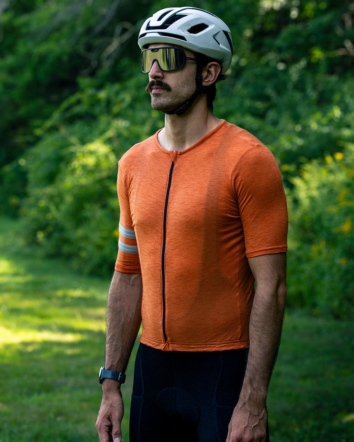 A man wearing a bright orange short-sleeve cycling jersey, black cycling shorts, a white cycling helmet, and reflective sunglasses, standing outdoors in a grassy area with trees in the background.