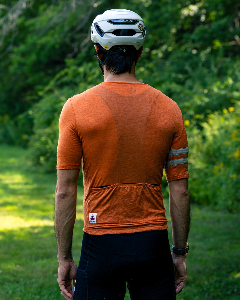 A man viewed from behind wearing a bright orange short-sleeve cycling jersey with reflective arm bands and black cycling shorts, standing outdoors on a grassy path with green trees in the background.