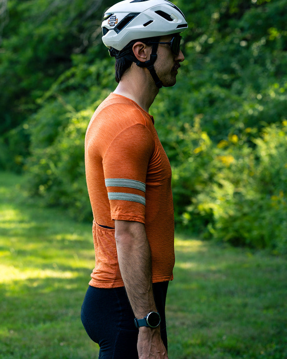 A man in a side profile wearing an orange short-sleeve cycling jersey with reflective stripes on the sleeve, black cycling shorts, and a white helmet, standing outdoors on a grassy path with green foliage in the background.