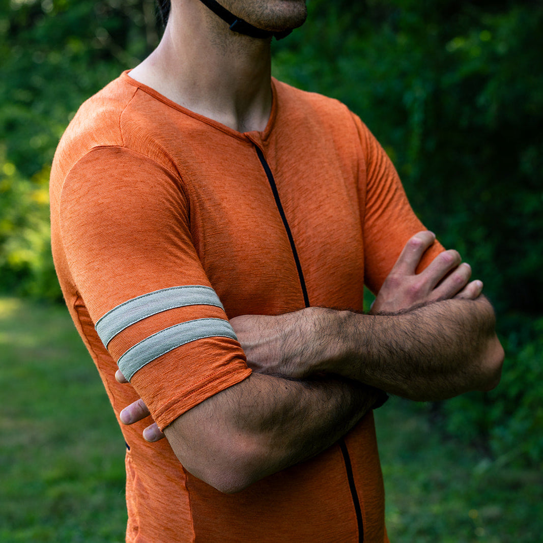 A man wearing an orange athletic long-sleeve shirt with gray sleeve stripes, standing outdoors with his arms crossed against a blurred green background.