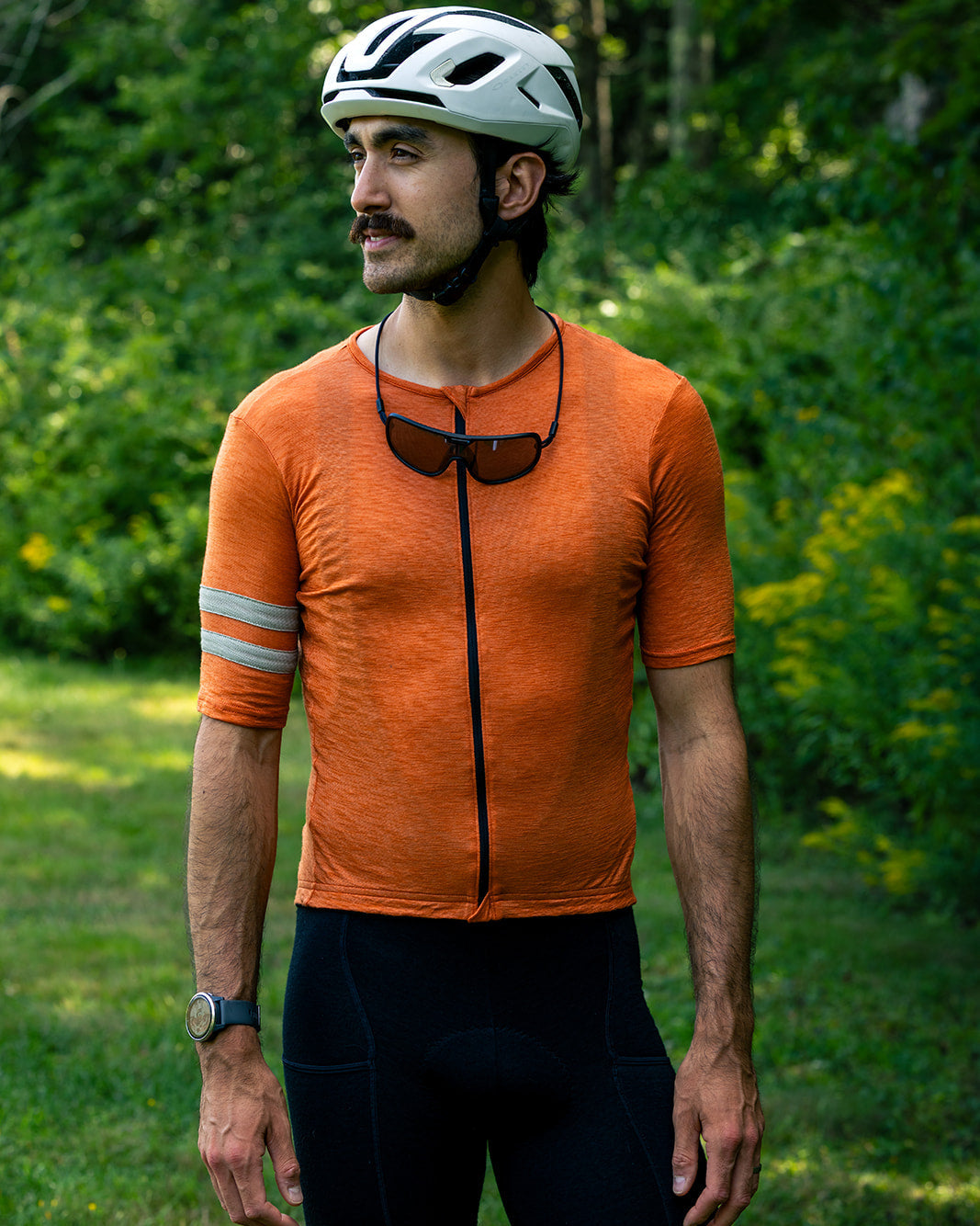 A man wearing a white bicycle helmet, an orange short-sleeve cycling jersey with a black front zipper and reflective arm tape, and black cycling shorts, standing outdoors in a grassy area with green trees.