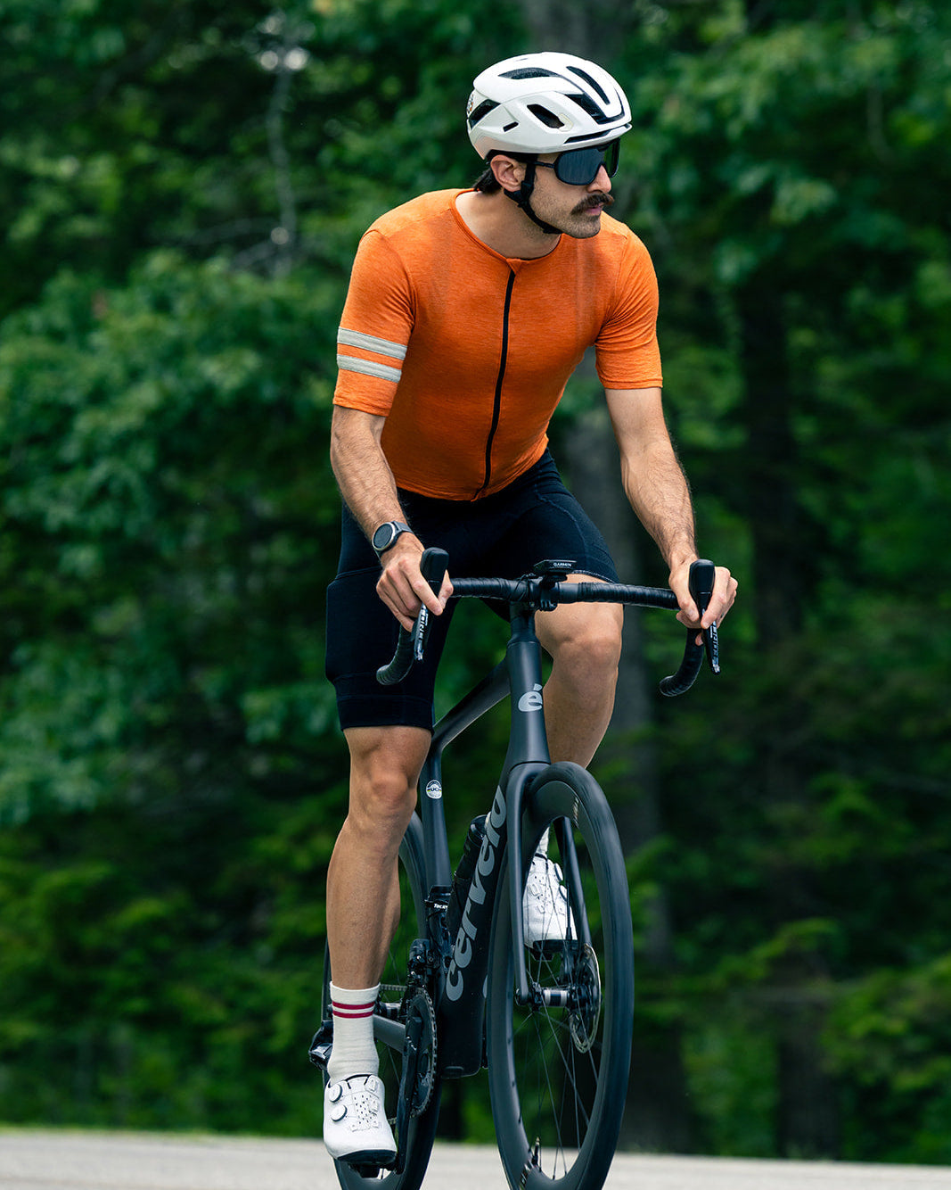 A man riding a black road bicycle outdoors on a paved path, wearing an orange short-sleeve cycling jersey with reflective stripes, black shorts, a white helmet, and sunglasses, with a blurred green forest background.