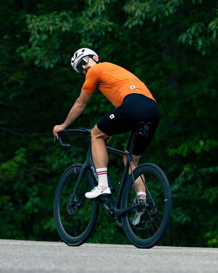 A cyclist wearing an orange jersey, black shorts, a white helmet, and white socks rides a black road bike on a paved path with a dense green forest in the background.