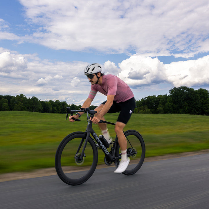 A man riding a black road bicycle on an asphalt road, wearing a pink jersey, black shorts, white socks, and a white helmet, with a green field and blue sky with clouds in the background.