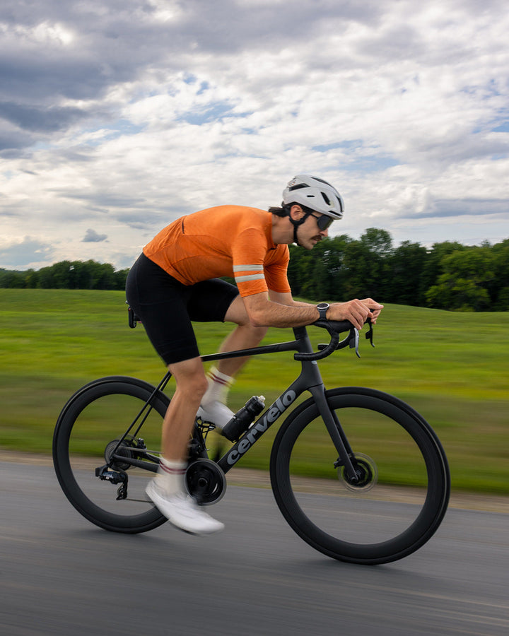 A cyclist in an orange jersey and black shorts rides a black road bike on a paved road with green fields and a cloudy sky in the background.