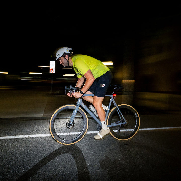 A cyclist riding a road bike at night on a dark road, wearing a bright neon yellow-green jersey, black shorts, and a white helmet. The background is heavily blurred due to motion, with streetlights creating horizontal light streaks.