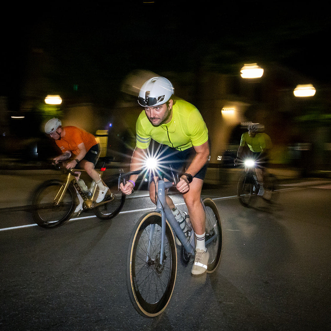 A cyclist in a bright neon yellow-green jersey and white helmet rides a road bike at night with a front light turned on, creating a bright flare on the pavement. Two other cyclists are visible in the blurred background on a dark street.
