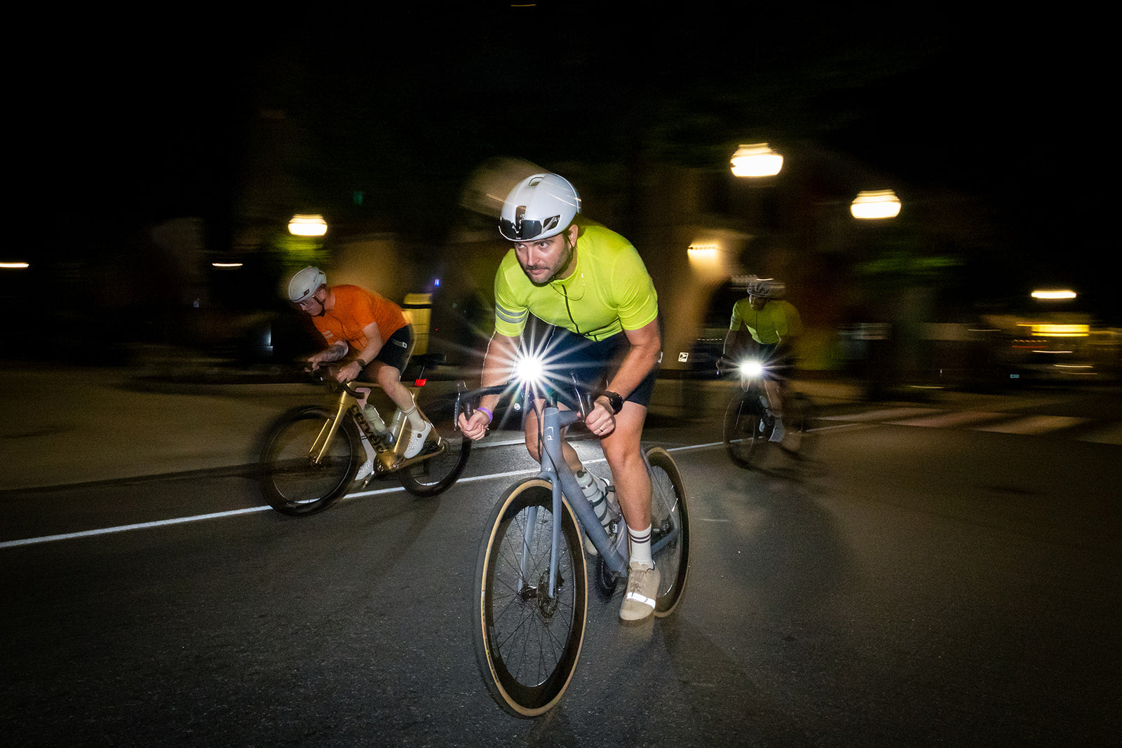 A cyclist in a bright neon yellow-green jersey and white helmet rides a road bike at night with a front light turned on, creating a bright flare on the pavement. Two other cyclists are visible in the blurred background on a dark street.
