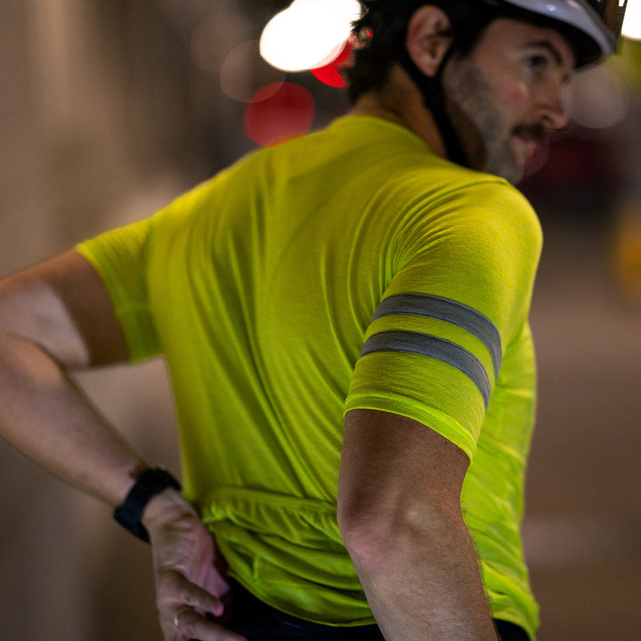A man wearing a bright neon yellow short-sleeve cycling jersey with reflective gray stripes on the sleeve, photographed from behind in low-light conditions.