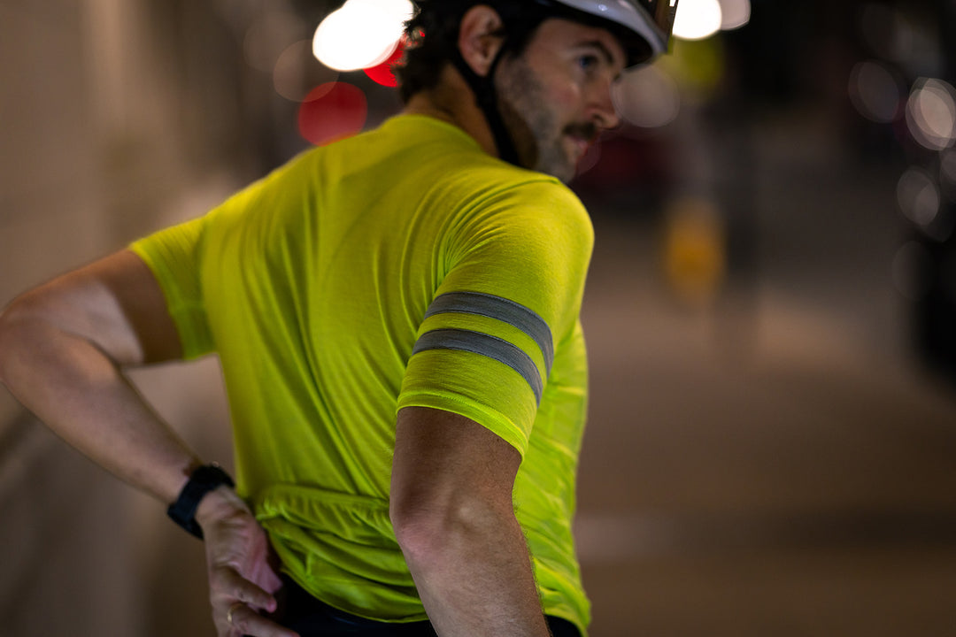 A man wearing a bright neon yellow short-sleeve cycling jersey with reflective gray stripes on the sleeve, photographed from behind in low-light conditions.