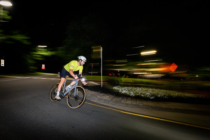 A cyclist in a bright yellow-green jersey and white helmet rides a road bike at night on a dark road, with motion blur and streetlights in the background.