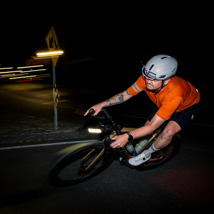 A cyclist wearing an orange jersey, black shorts, and a white helmet rides a bicycle at night on a dark road, with the bicycle headlight illuminating the path.