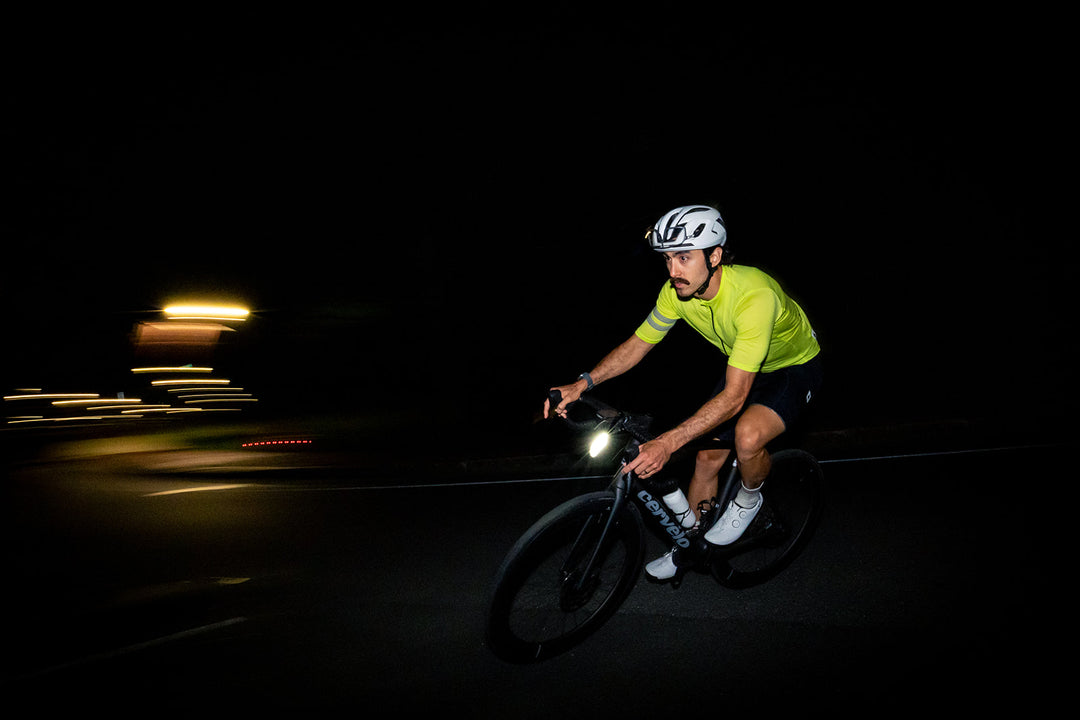 A cyclist riding at night wearing a bright neon yellow-green cycling jersey, black shorts, white cycling shoes, and a white helmet. The cyclist is riding a dark road bike with a headlight on, set against a dark road with blurred streetlights on the left.