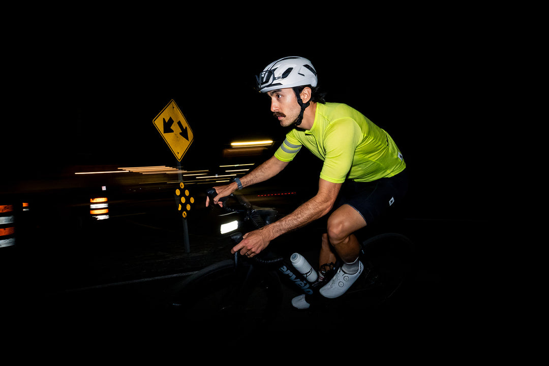 A cyclist riding at night wearing a bright neon yellow-green jersey, black shorts, white cycling shoes, and a white helmet. The cyclist is illuminated by a headlight on a dark road with a yellow road sign in the background.