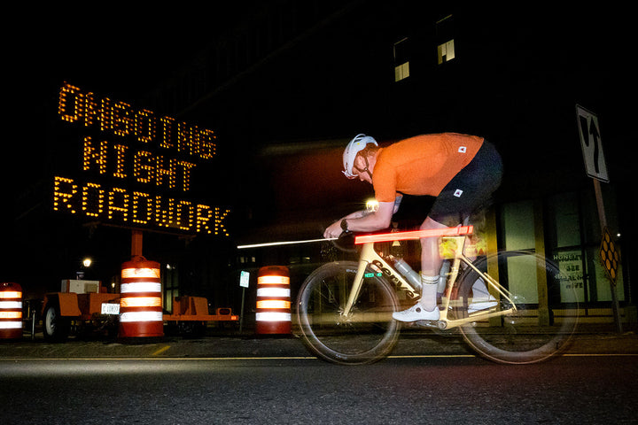 A cyclist in an orange jersey and black shorts rides a road bike at night on a city street, with a large illuminated &
