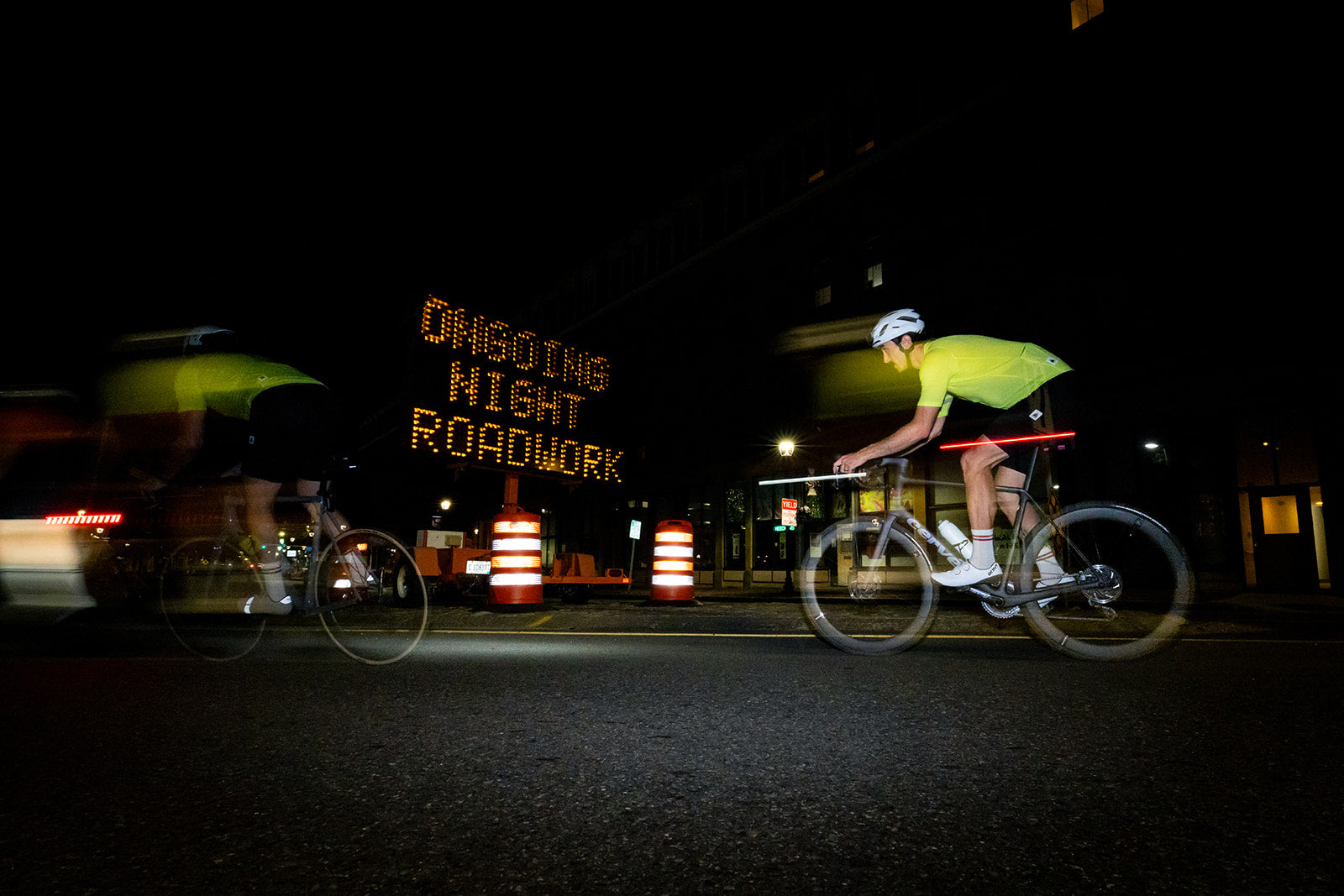 A cyclist in a bright neon yellow jersey rides a road bike at night near a large illuminated sign reading 'ONGOING NIGHT ROADWORK'. Another cyclist is blurred in motion on the left, and traffic cones are visible on the street.