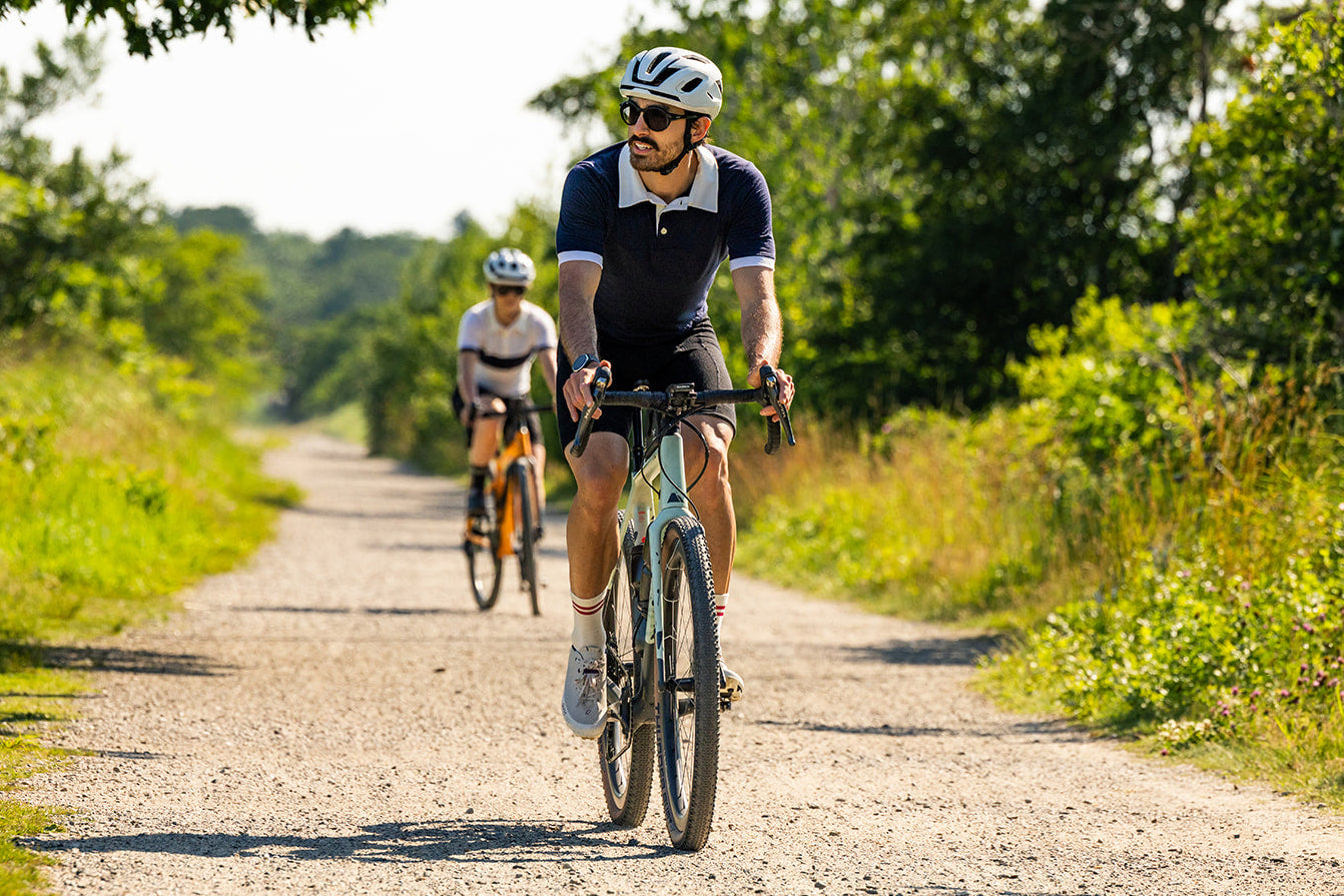 A cyclist wearing a navy and white cycling jersey, white helmet, and sunglasses rides on a sunlit dirt path, with another rider visible in the background among green trees and grass.