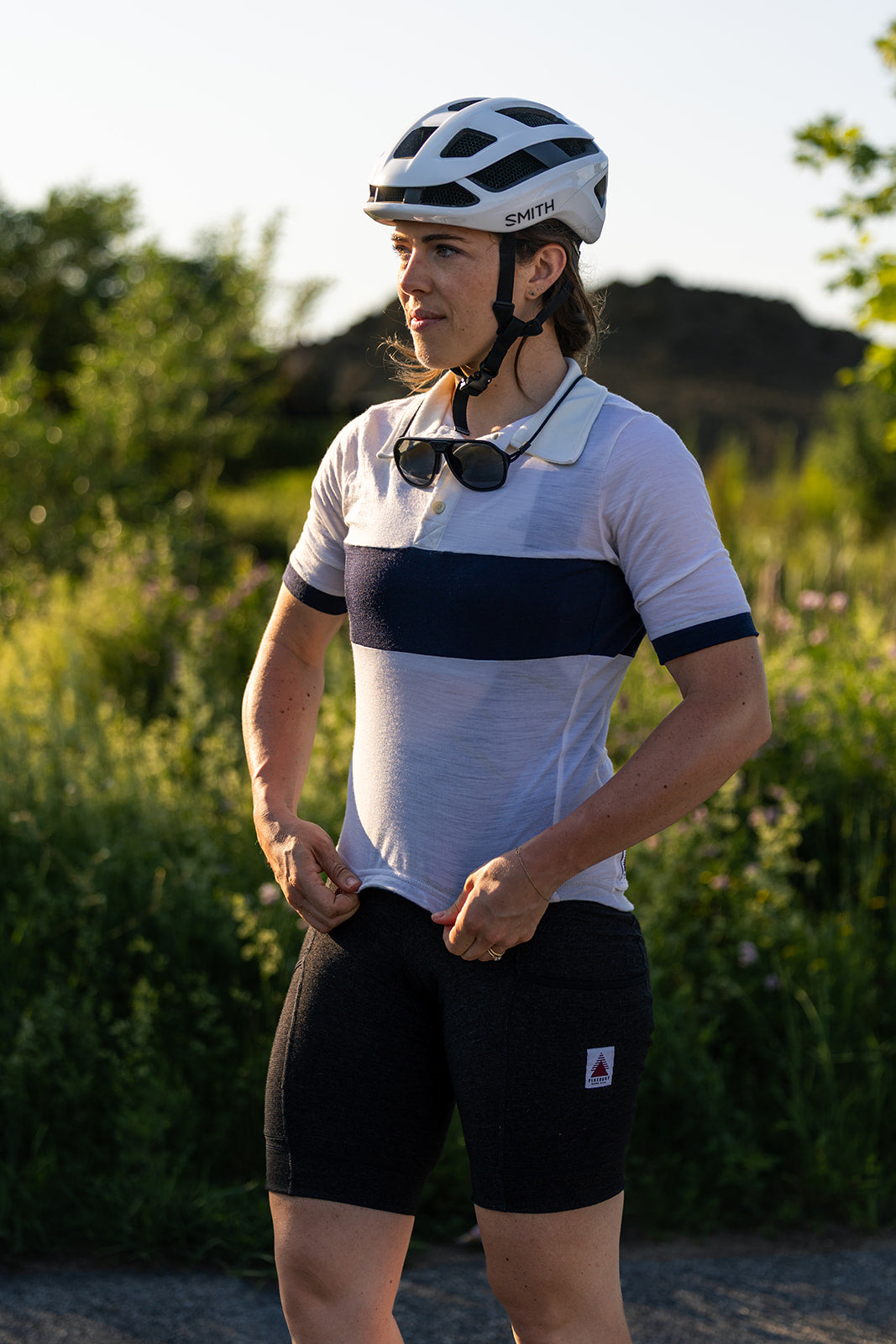 A cyclist wearing a white and navy blue short-sleeve jersey, black shorts, and a white helmet stands outdoors in a grassy field.
