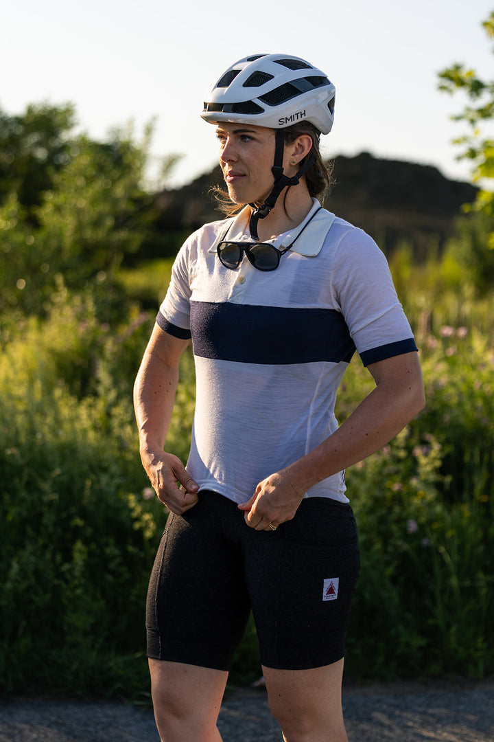 A cyclist wearing a white and navy blue short-sleeve jersey, black shorts, and a white helmet stands outdoors in a grassy field.
