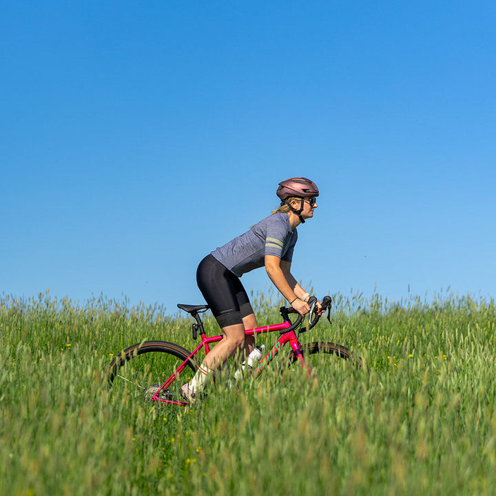 A cyclist wearing a pink helmet, gray jersey, and black shorts rides a pink bicycle across a green grassy field under a clear blue sky.