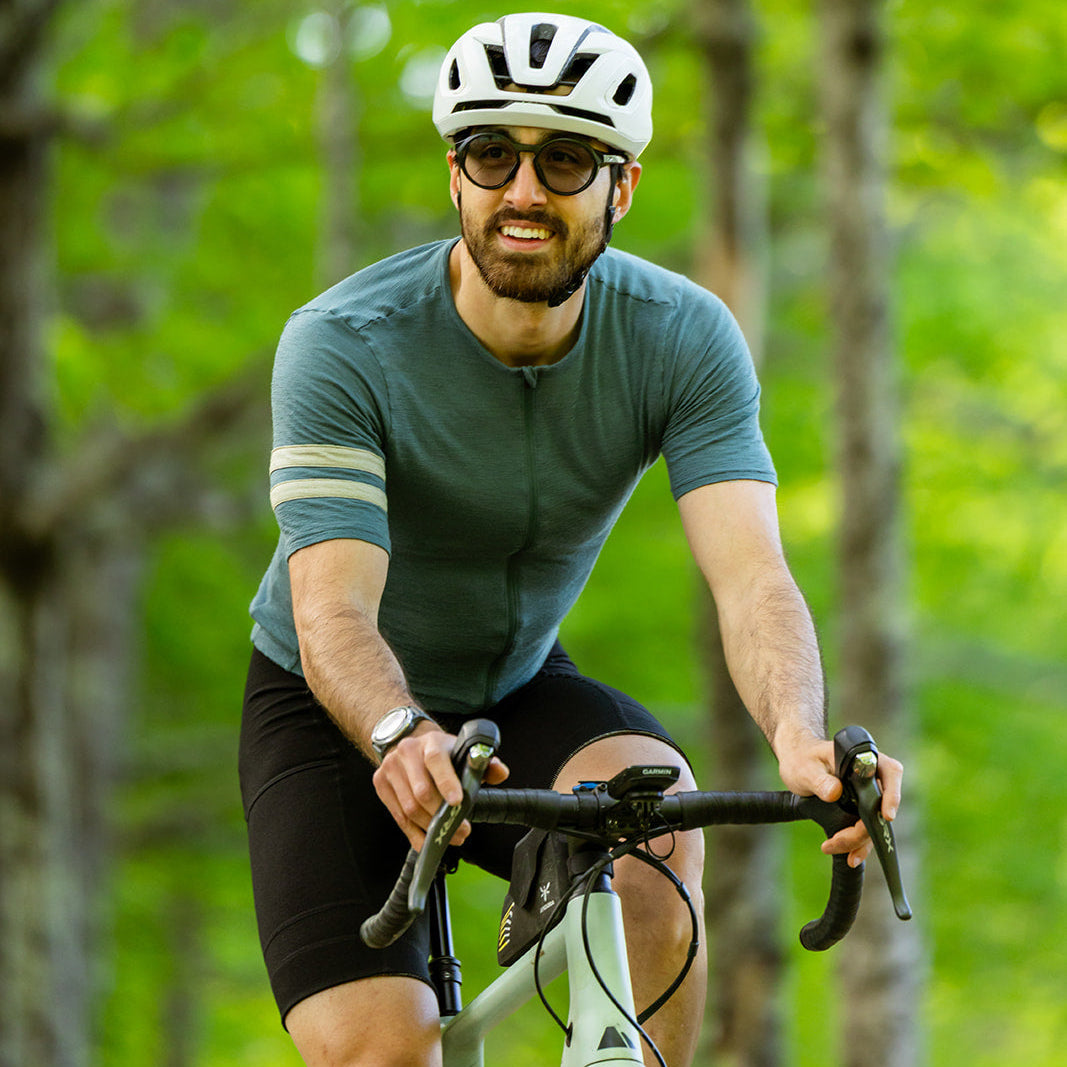 A man wearing a white bicycle helmet, sunglasses, a teal short-sleeve cycling jersey, and black shorts rides a light-colored bicycle on a forest path with green trees in the background.