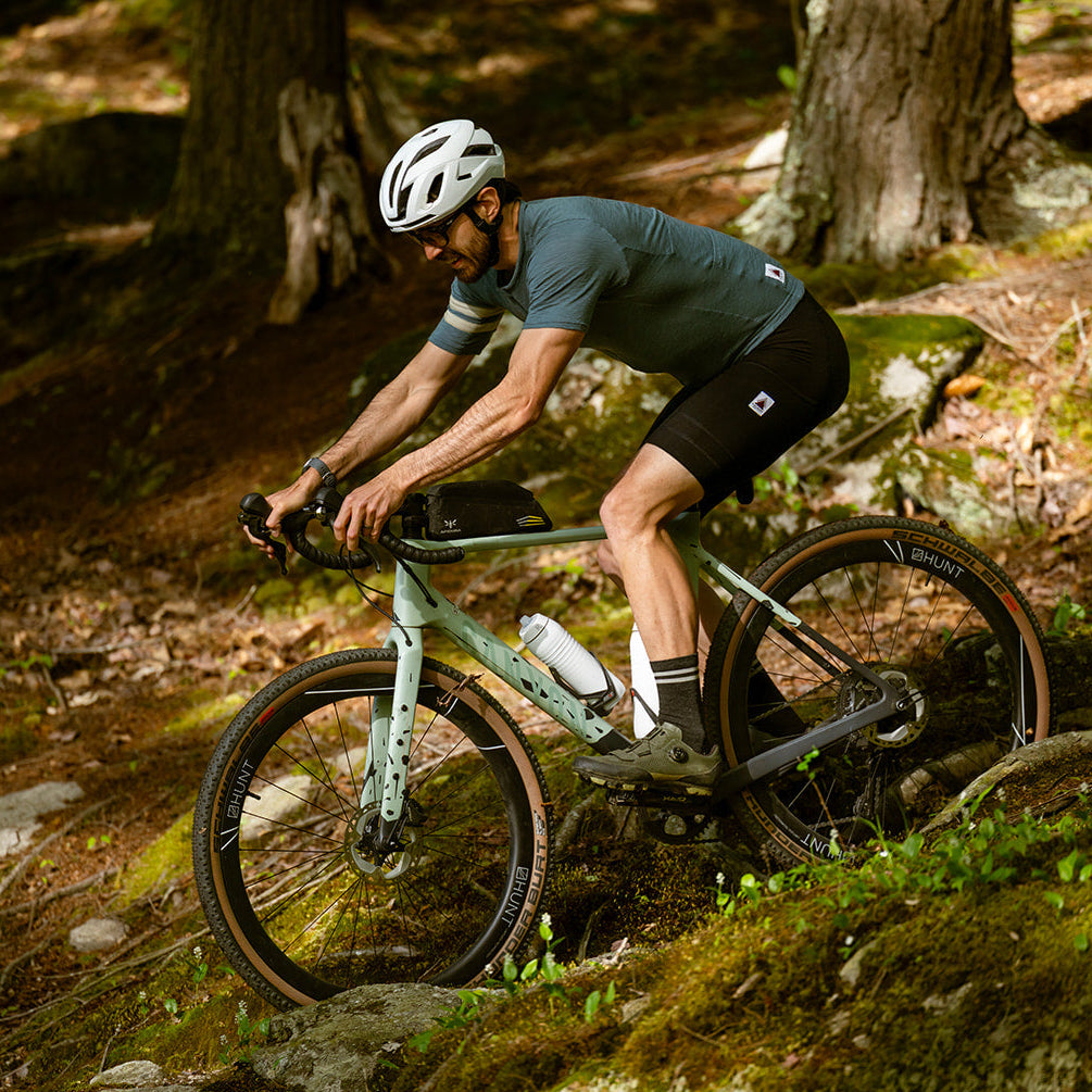 A cyclist wearing a white helmet, dark green jersey, and black shorts rides a gravel bike through a rocky, mossy forest trail.
