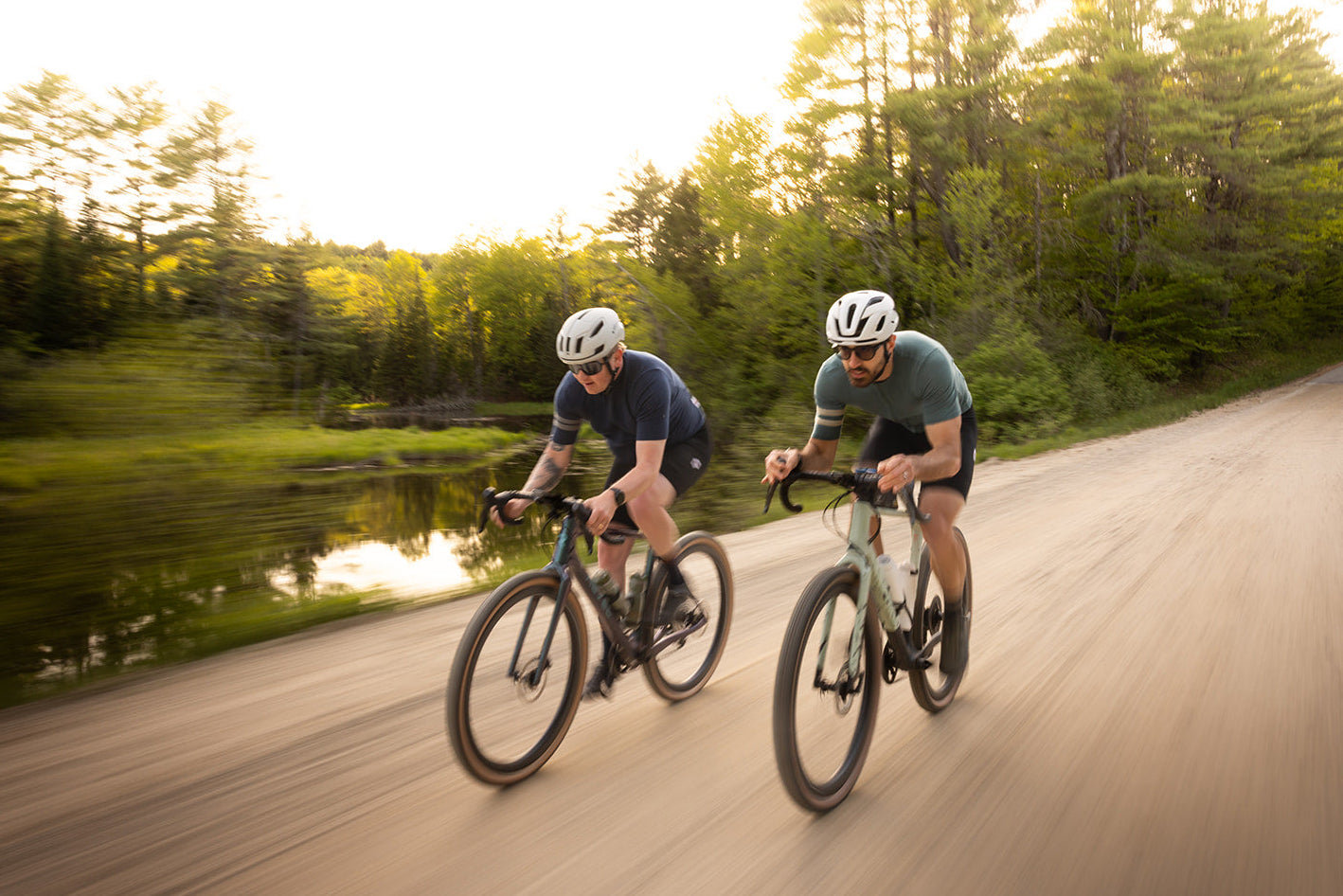 Two cyclists riding on a paved road surrounded by trees and a lake.