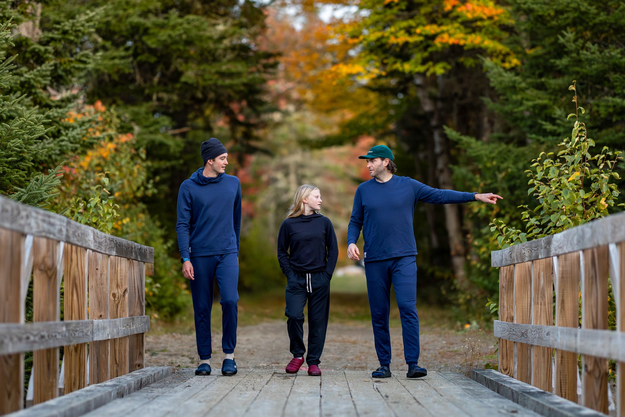 Three people walking on a wooden bridge in a forest with autumn foliage.
