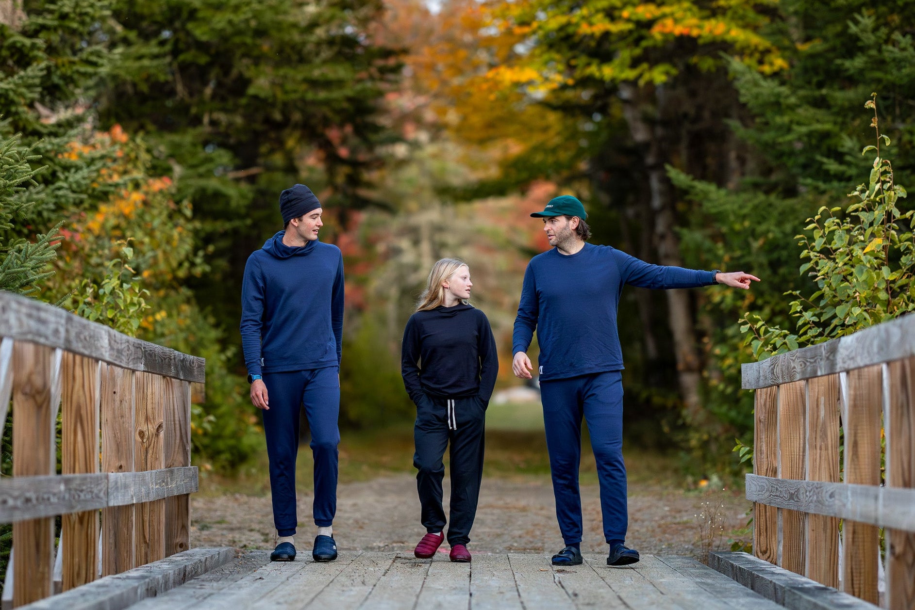 Three people walking on a wooden bridge in a forest with autumn foliage.