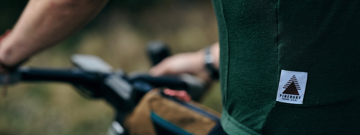 A close up show of the logo details on the back pocket of a Pinebury Rangeley Short Sleeve Merino Wool Jersey in Pine with an out of focus bikes handlebars in the background.